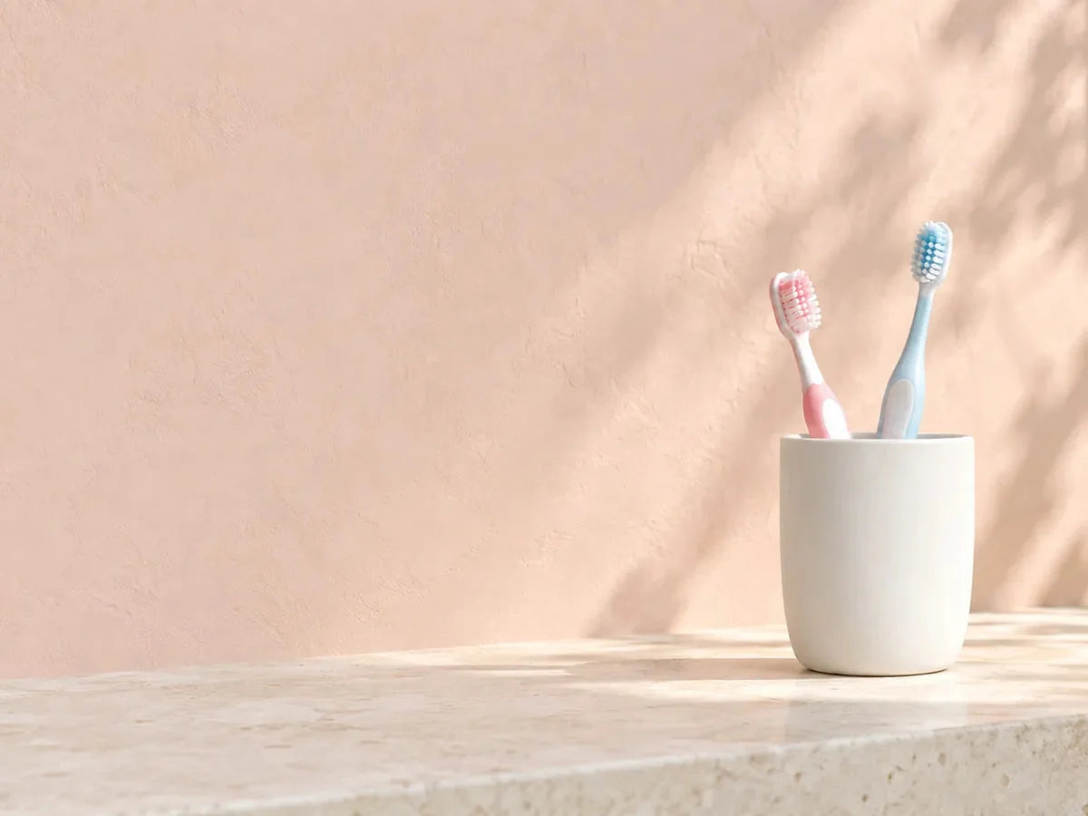 A pink and a blue toothbrush together in a cup on a sunny counter